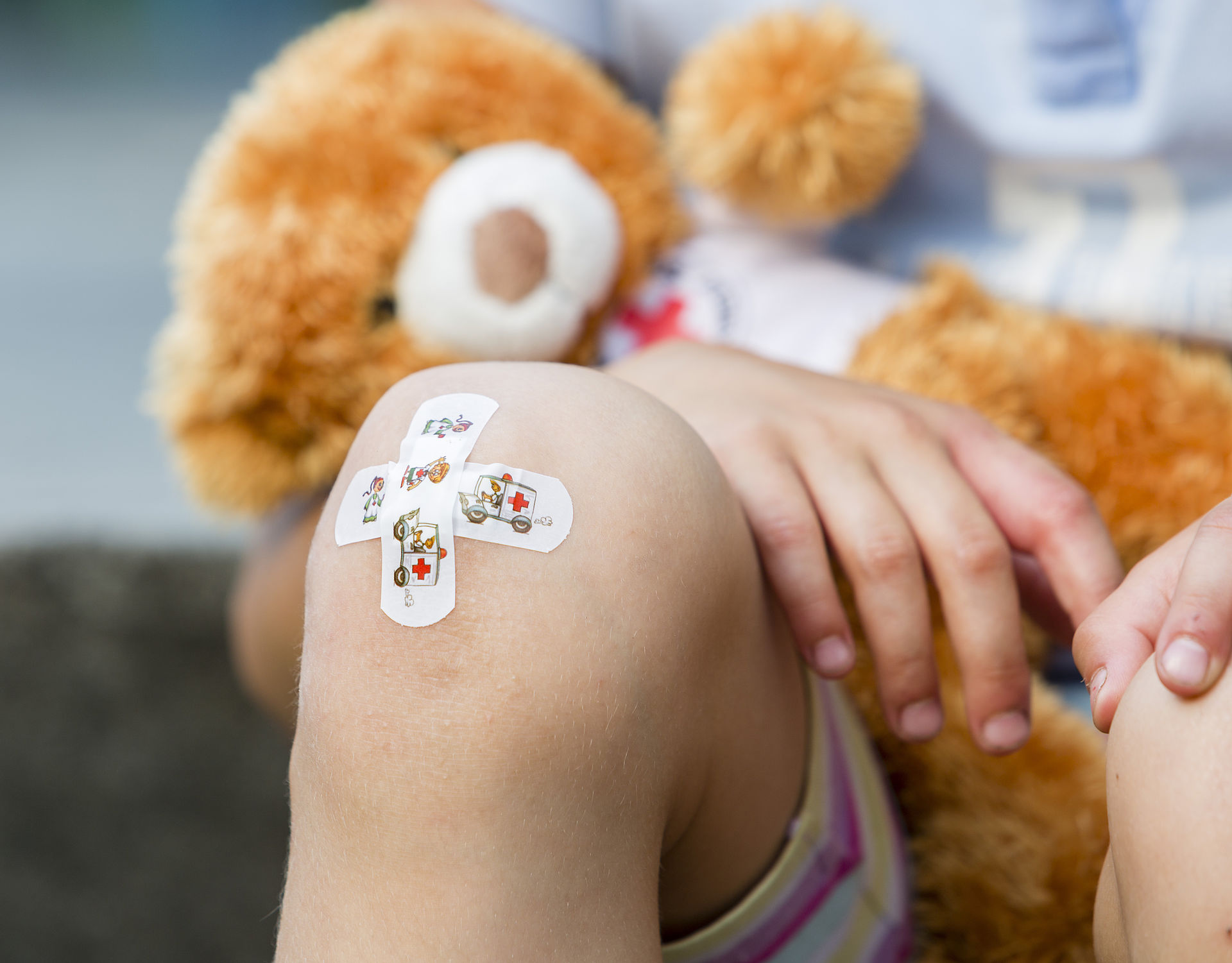 Ein Kind hat ein Teddybär im Arm und ein Pflaster am Knie.